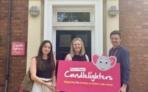 2 women and a man standing in front of a black front door holding a pink sign with the Candlelighters trust logo on